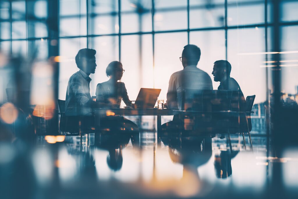 Four business leaders meeting in a glass-walled conference room, seated around a table with a laptop, collaborating on strategy and discussing coordinated risk management for a growing mid-market organization.