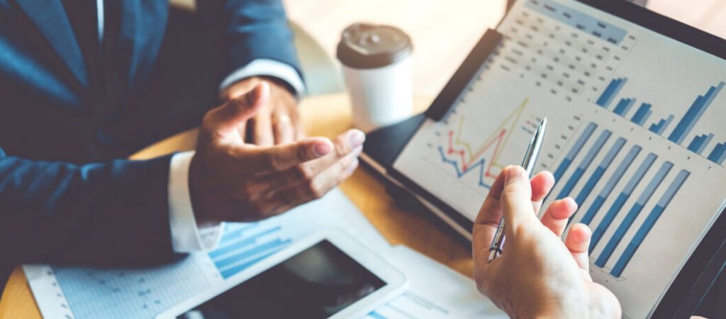 Two professionals pointing and consulting graphs on a tablet and data on paperwork. Their meeting represents the partnership between Optimal and financial teams to reach desired levels of financial control.
